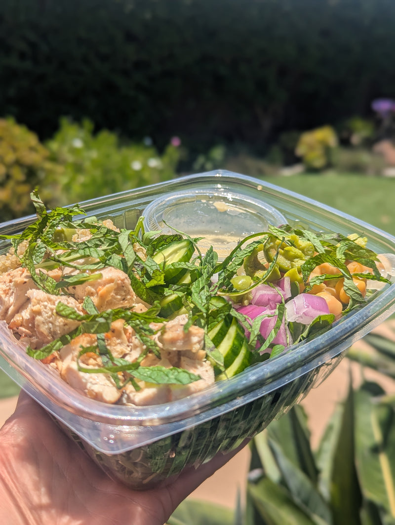 Container of salad with greens and possibly chicken or tofu held outdoors.